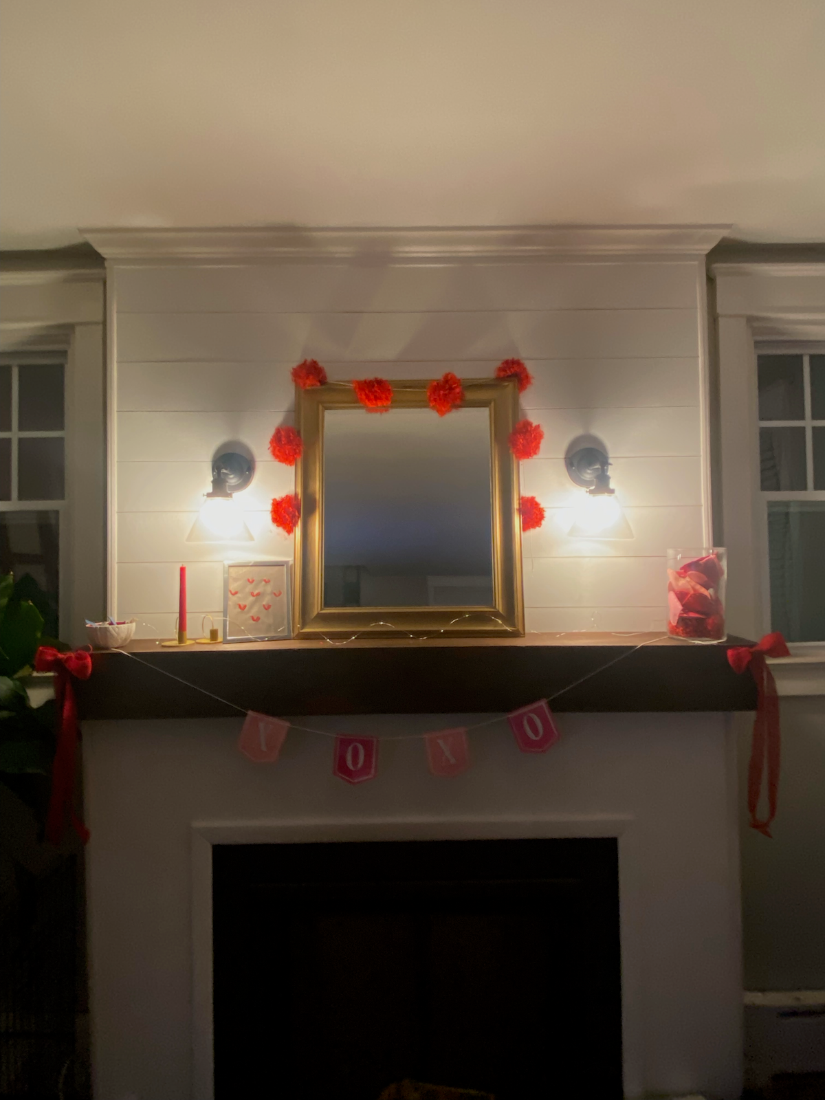 Cozy Valentine’s Day mantel featuring a gold-framed mirror outlined with red pom-poms, soft wall lighting, candles, and a pink XOXO banner above a white fireplace.